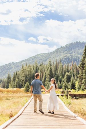 Couple holding hands while walking on a wooden boardwalk through a mountain meadow with pine trees and clouds.