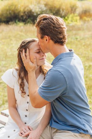 Man tenderly kissing woman on forehead while gently holding her face in a sunlit outdoor setting.