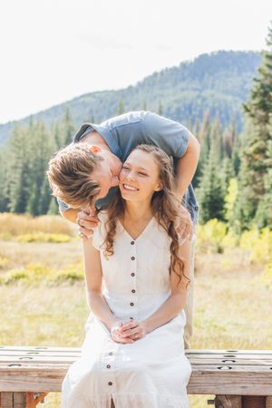 Man leaning over woman seated on wooden fence, kissing her cheek, mountain forest backdrop.