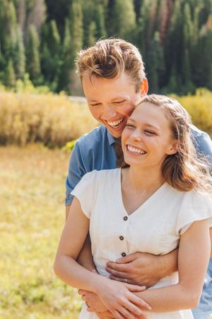Happy couple embracing outdoors, laughing together in a sunny meadow with green trees in the background.