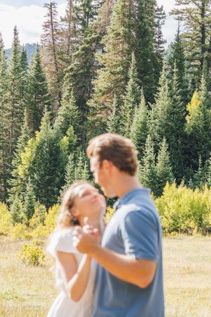 Couple dancing together in a sunny mountain meadow surrounded by tall evergreen trees and golden foliage.