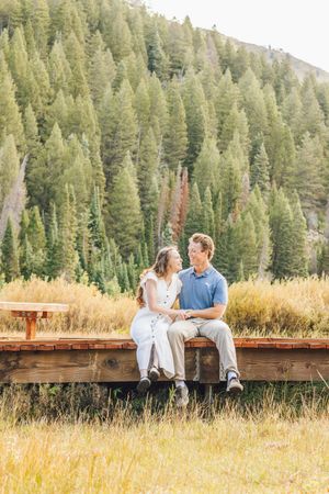 Couple sitting on a wooden fence in a mountain meadow, surrounded by tall pine trees and golden grass.