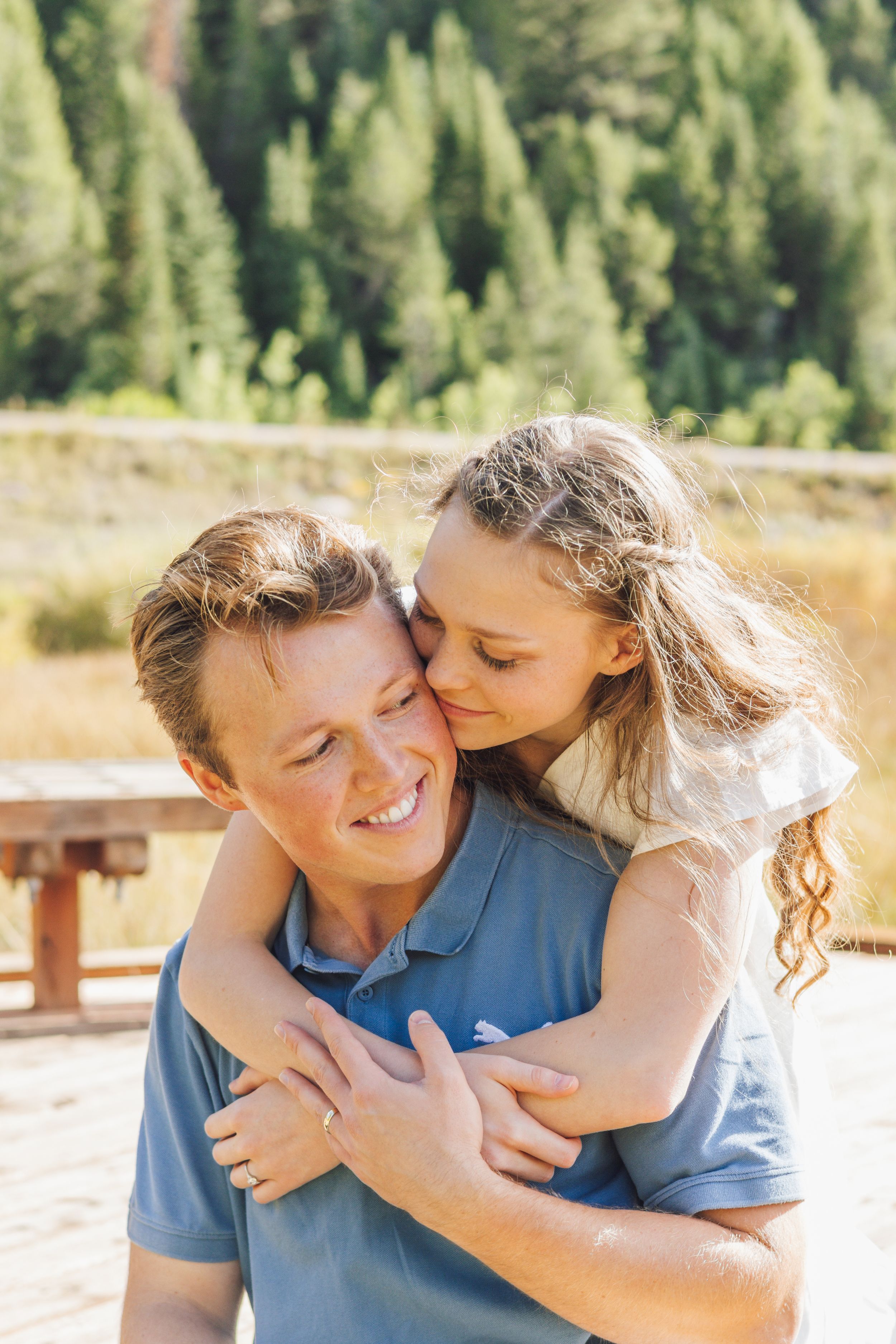 Smiling man gives piggyback ride to curly-haired girl kissing his cheek outdoors near pine forest.
