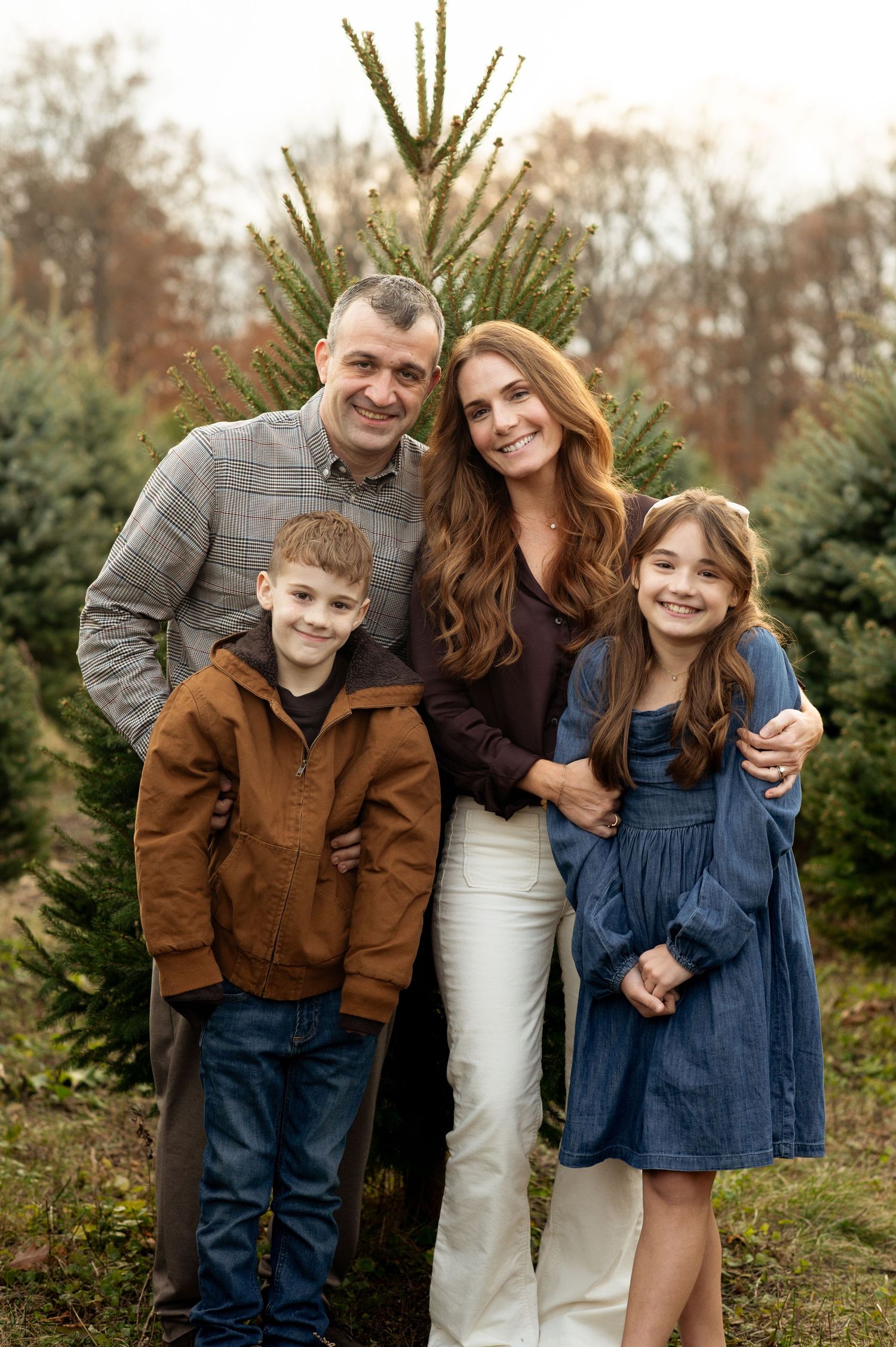 Family of four posing together at a Christmas tree farm, parents with two children smiling outdoors.