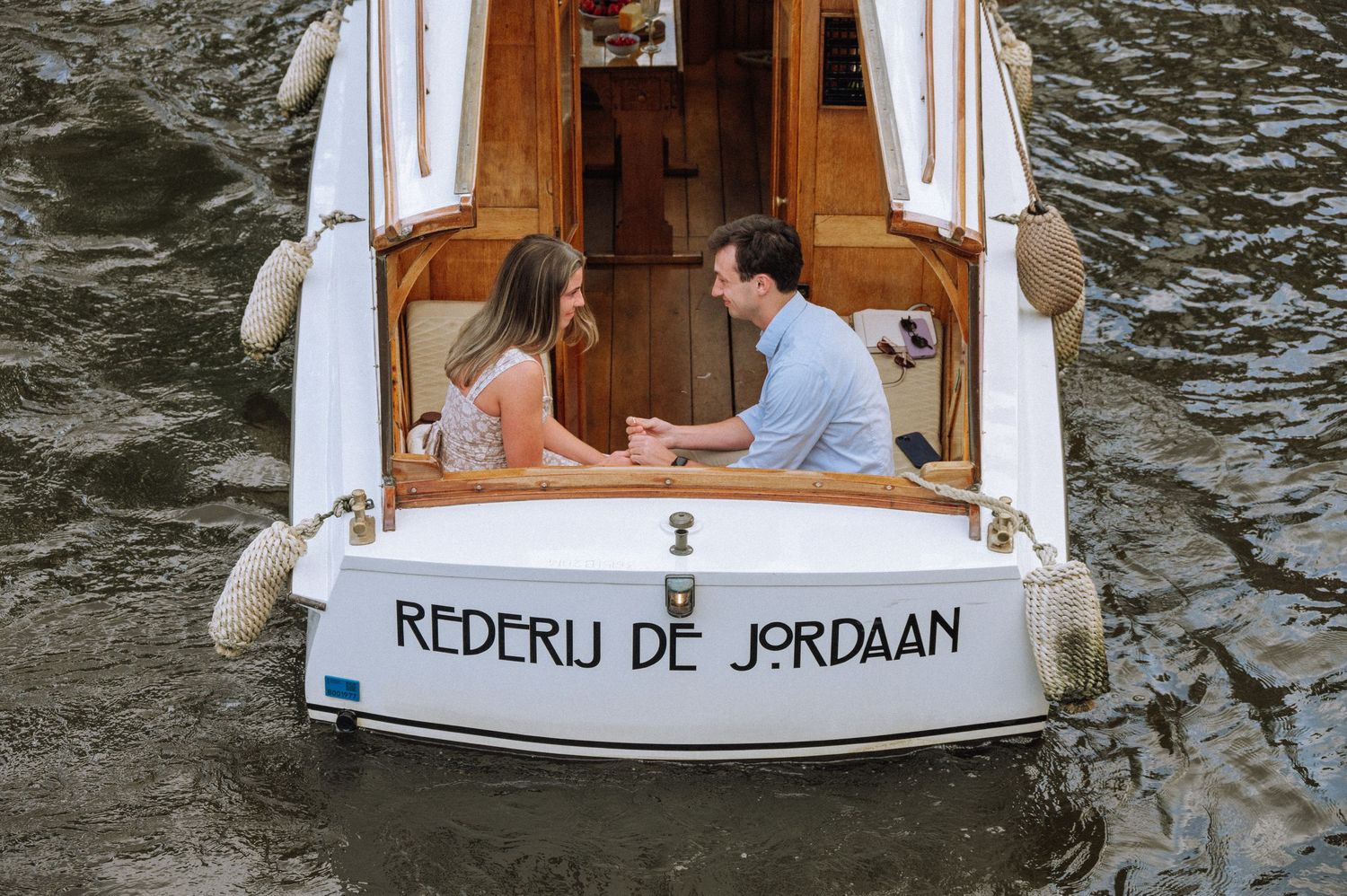 Couple sharing a romantic kiss on a wooden boat along a scenic Amsterdam canal with a stone bridge backdrop.