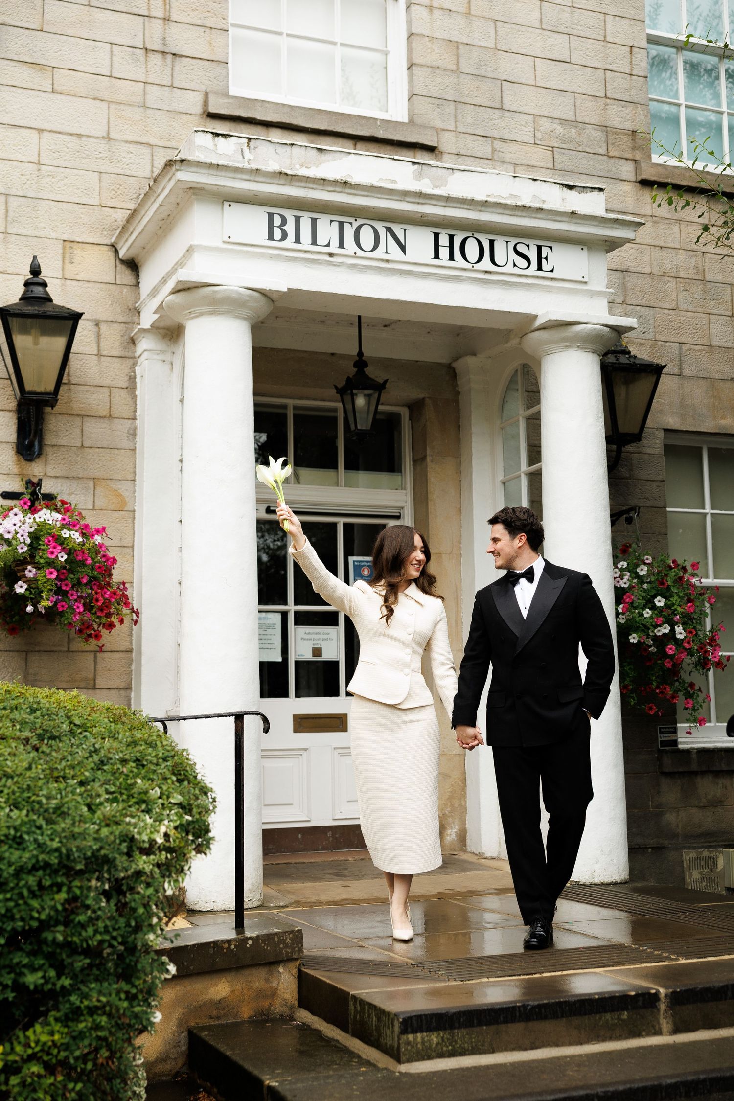 Elegant couple in wedding attire walking out of Bilton House, bride in white suit holding flowers, groom in black tuxedo.