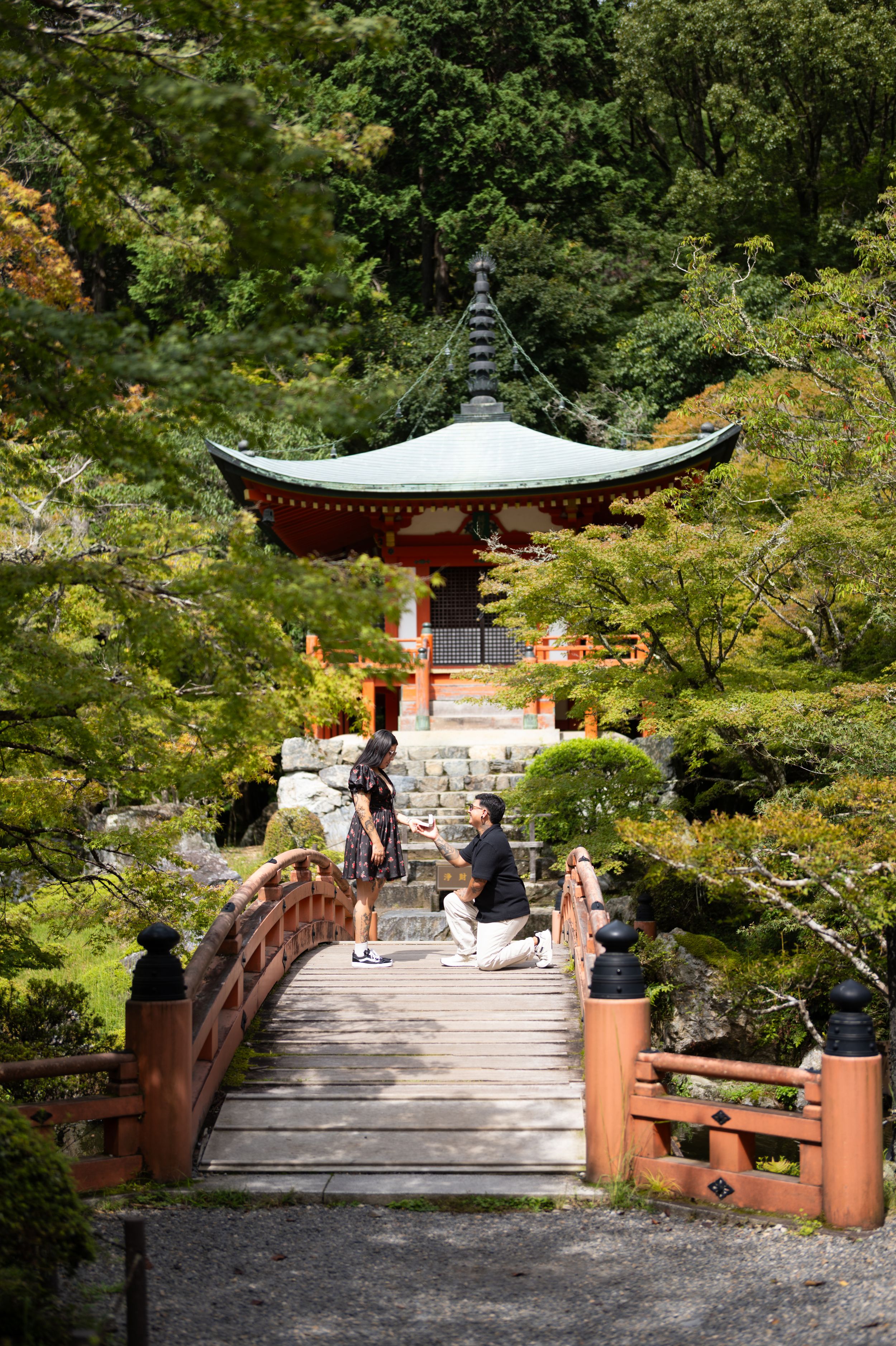 Couple standing on a wooden bridge surrounded by lush green trees in a Japanese garden.