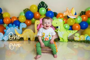 Baby boy smiles at his first birthday dinosaur themed cake smash with colorful balloons and dino decorations.
