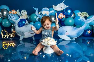 Baby smashing first birthday cake surrounded by ocean-themed balloons including sharks and whales on a blue backdrop.