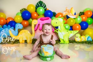 Baby smiling at first birthday cake smash with colorful dinosaur decorations and balloon garland backdrop.