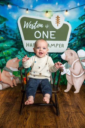 Baby Weston sits in a wooden chair smiling at his first birthday camping themed photo shoot.