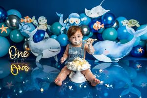 Baby smashing first birthday cake surrounded by ocean-themed balloons and sea creature decorations on a blue backdrop.