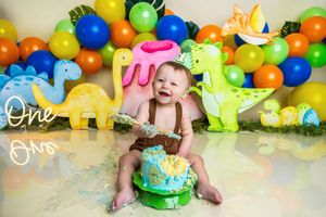 Baby boy smashing first birthday cake at dinosaur themed party with colorful balloons and dino decorations.