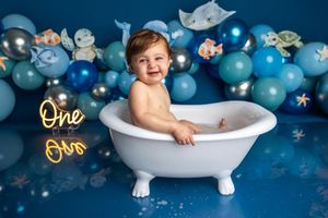 Baby boy smiling in white bathtub surrounded by blue balloons and ocean decorations at a first birthday cake smash.