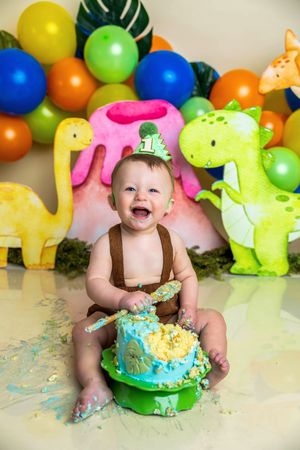 Baby boy smashing dinosaur-themed first birthday cake, laughing joyfully, surrounded by colorful balloons and dino decorations.