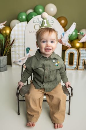 Smiling baby boy in green shirt and party hat celebrates his first birthday with balloon decorations.