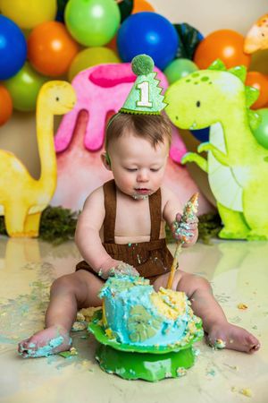 Baby's first birthday cake smash with dinosaur theme, colorful balloons, and a number one party hat.
