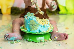 Baby smashing a blue and green first birthday cake, with crumbs scattered on the floor during a cake smash photoshoot.