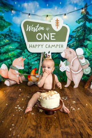 One-year-old Weston enjoys a happy camper cake smash surrounded by cute forest animal cutouts and a festive sign.