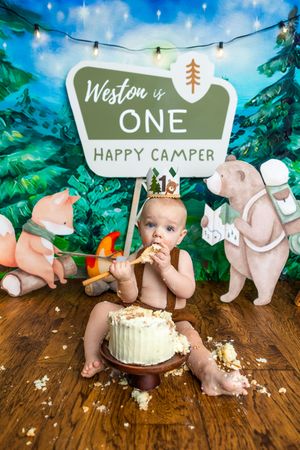 Baby Weston celebrates his first birthday with a camping-themed cake smash, wearing a crown amid woodland animal decorations.