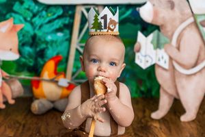 Baby in a crown celebrating first birthday, eating cake smash treat with woodland animal decorations in background.