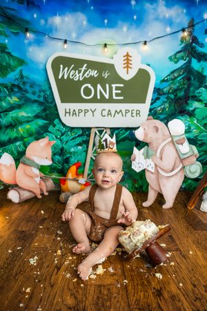 Baby Weston celebrates his first birthday with a camping-themed cake smash, surrounded by bear figurines and a Happy Camper sign.