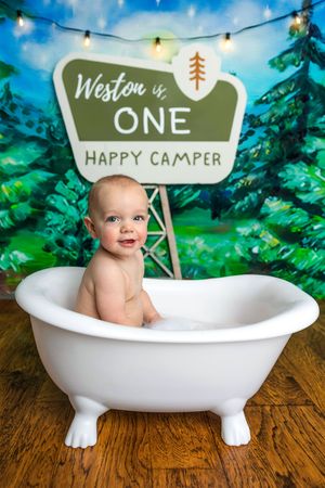 Baby boy sits smiling in a white clawfoot tub in front of a camping-themed backdrop celebrating Weston turning one.