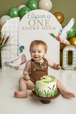 Baby Ellyson celebrates first birthday cake smash with duck theme, green balloons, and decorated cake.