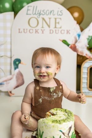 Baby Ellyson celebrates turning ONE at a duck-themed cake smash birthday party with green frosting on face.