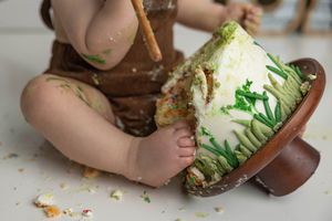 Baby smashing a dinosaur egg-shaped first birthday cake, covered in green frosting decorations.