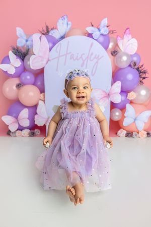 Smiling baby girl Haisley in sparkly lavender dress at butterfly balloon first birthday celebration backdrop.
