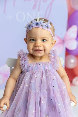 Smiling baby girl in purple tulle dress and floral headband at her first birthday party celebration.
