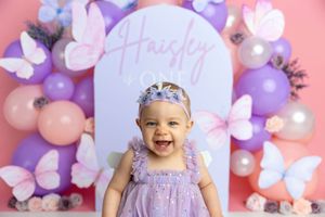 Smiling baby girl in purple dress celebrates first birthday with butterfly and balloon backdrop spelling Haisley ONe.