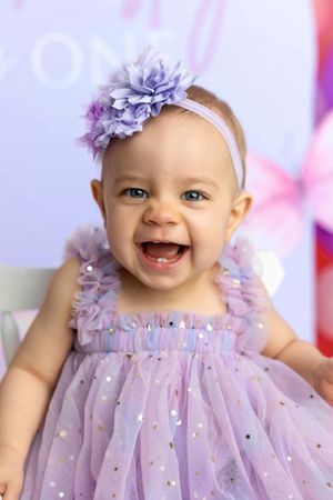 A smiling baby girl in a purple tulle dress with sequins and a floral headband celebrates her first birthday.