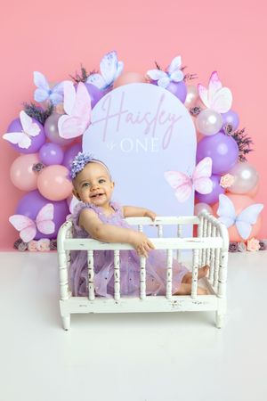 Smiling baby girl in purple dress sits in white crib at butterfly-themed first birthday party with balloon backdrop.