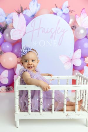 Baby girl Haisley in purple tulle dress sitting in white crib at butterfly-themed first birthday photo shoot.
