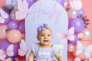 A smiling baby girl in a purple outfit sits before a butterfly-themed first birthday backdrop with balloons spelling Haisley is ONE.