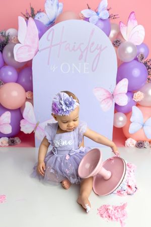 Baby girl in purple tutu celebrates first birthday with butterfly balloon backdrop and smash cake.