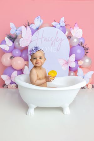 Baby girl named Haisley sits in white clawfoot tub holding yellow duck, surrounded by purple pink balloons and butterflies.