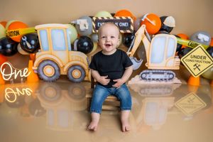 Baby boy smiles in black shirt and jeans surrounded by construction-themed balloons and signs for his first birthday photo.