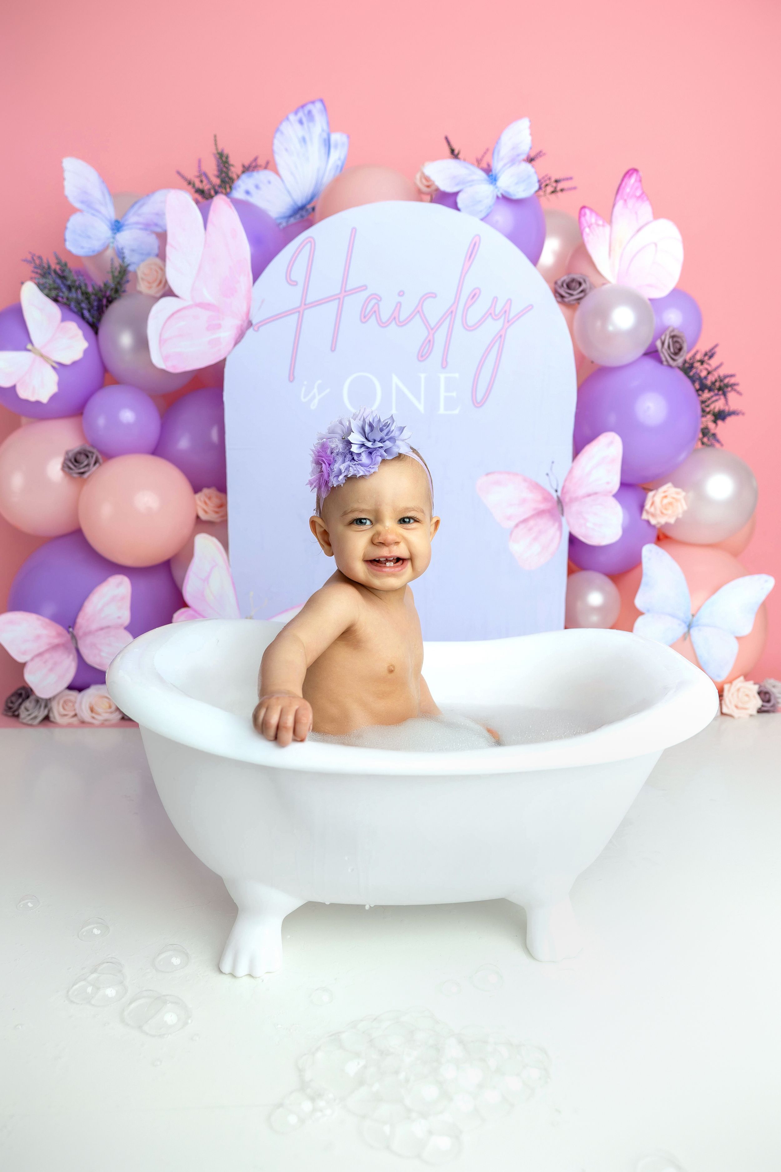 Baby girl Haisley sits in a white bathtub surrounded by purple balloons and butterflies for her first birthday.