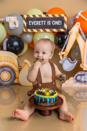 Baby Everett celebrates his first birthday with a construction-themed cake smash, surrounded by trucks and colorful balloons.