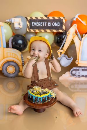 Baby Everett celebrates his first birthday with a construction-themed cake smash, wearing a yellow hard hat and brown overalls.
