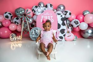 Baby girl sits in white chair at pink cow-themed first birthday party with balloons, disco balls, and guitar backdrop.
