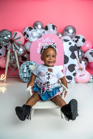 Baby girl sits on white bench at pink cowgirl first birthday party with balloons and guitar backdrop.
