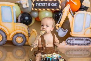 Baby boy in brown suspenders smashes first birthday cake at construction-themed party with excavator decorations.