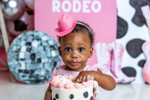 A baby girl in a pink cowboy hat touches her first birthday cake at a rodeo-themed party with disco balls.