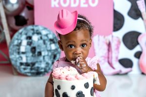 Baby girl in pink cowboy hat smashes first birthday cake at rodeo-themed party with disco ball decor.