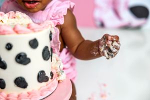 Baby girl in pink ruffle dress smashing first birthday cake with black spots and pink frosting decorations.