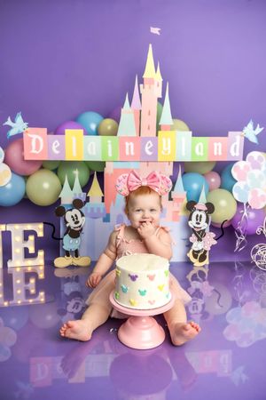 Baby girl in pink bow sits before a Disney-themed first birthday cake smash with Cinderella castle backdrop.